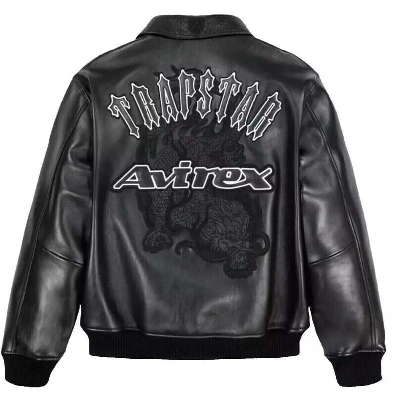 May include: Black leather jacket with a bomber-style design. The back features the text "TRAPSTAR" above a dragon graphic and the "Avirex" logo in white. The jacket has a collar, ribbed cuffs, and a ribbed hem.