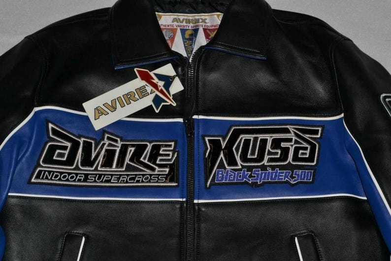 Puede incluir: Chaqueta de cuero negra con detalles azules y blancos. Presenta la marca "AVIREX" y los textos "INDOOR SUPERCROSS" y "Black Spider 500". Incluye cierre de cremallera y etiqueta.