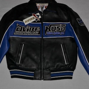 Puede incluir: Chaqueta de cuero negra con detalles azules y blancos. Presenta el texto "AVIRE Indoor Supercross" y "XUSA Black Spider 500". Incluye cierre de cremallera, bolsillos y pu&ntilde;os y dobladillo acanalados.