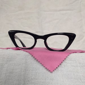 May include: Black cat-eye glasses with a pink cleaning cloth. The glasses have a thin black frame and clear lenses.