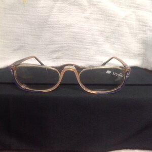 May include: Clear, brown-rimmed glasses with blue accents. The glasses have a studio logo on the right lens.