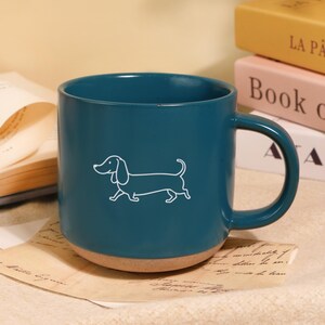 Puede incluir: Taza de cerámica color turquesa con una ilustración de perro salchicha blanco. La taza tiene forma redondeada, un asa robusta y una base de color natural. La taza está sobre un papel manuscrito, con libros al fondo.