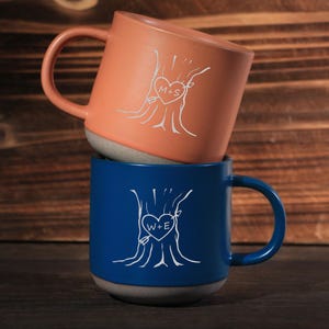 Op de afbeelding: Twee keramische mokken, één koraal en één blauw, op elkaar gestapeld. Elke mok heeft een wit boomontwerp met een hart en initialen in de stam gegraveerd. De koraal mok heeft de initialen "M+S", en de blauwe mok "W+E".