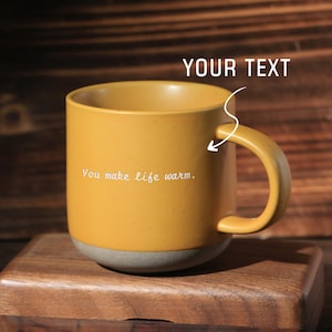 May include: Mustard yellow ceramic mug with a gray base and a curved handle. The mug has the text "You make life warm." printed on the side. The mug is sitting on a wooden surface.