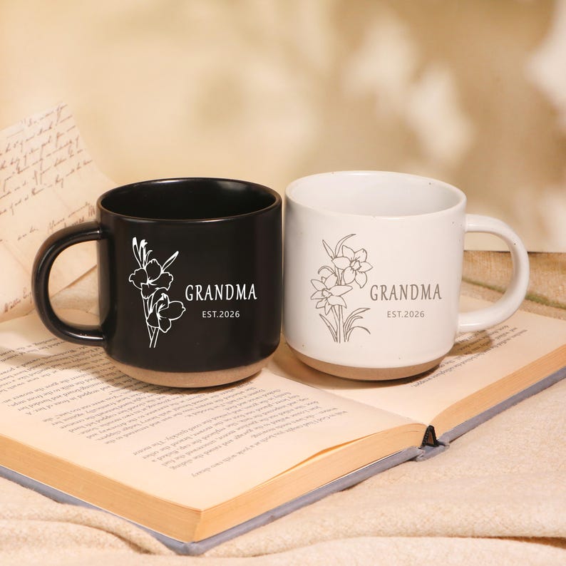 Peut inclure: Deux mugs en c&eacute;ramique, un noir et un blanc, chacun avec un motif floral et l'inscription "GRANDMA" et "EST.2026". Les mugs sont pos&eacute;s sur un livre ouvert.