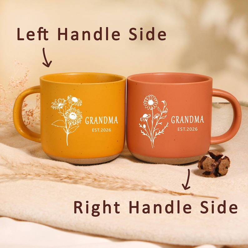 Peut inclure: Deux mugs en c&eacute;ramique, un jaune moutarde et un terracotta, avec des motifs floraux blancs. Chaque mug porte l'inscription "GRANDMA" et "EST.2026". Les mugs sont pos&eacute;s sur une surface aux tons neutres.