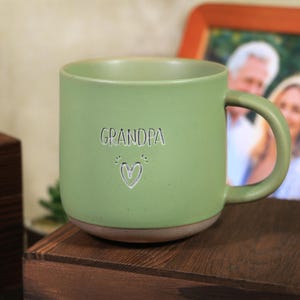 Könnte beinhalten: Eine grüne Keramik-Tasse mit dem Wort "GRANDPA" und einem Herz-Design. Die Tasse hat eine abgerundete Form, einen Henkel und einen hellbraunen Boden. Die Tasse steht auf einer Holzoberfläche.