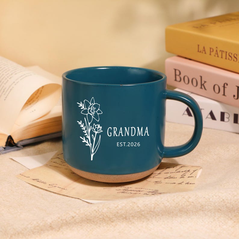 Peut inclure: Mug en c&eacute;ramique bleu sarcelle avec un motif floral blanc et le mot "GRANDMA". La base du mug est marron clair et le texte "EST.2026". Le mug est pos&eacute; sur un papier avec un livre ouvert et d'autres livres en arri&egrave;re-plan.