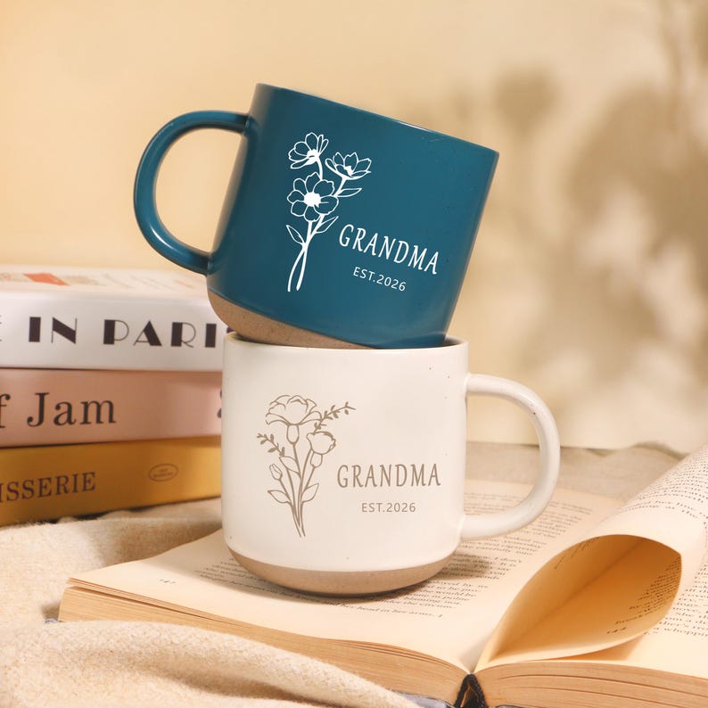 Peut inclure: Deux mugs en c&eacute;ramique, un bleu canard et un blanc, empil&eacute;s. Chaque mug pr&eacute;sente un motif floral et le mot "GRANDMA" avec "EST.2026". Les mugs ont une base naturelle non &eacute;maill&eacute;e et une anse incurv&eacute;e.