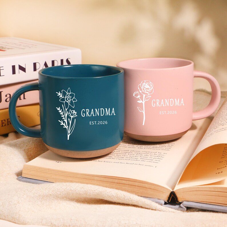 Peut inclure: Deux mugs en c&eacute;ramique, un bleu canard et un rose, avec des motifs floraux et le mot "GRANDMA" en blanc. Le mug bleu canard pr&eacute;sente une jonquille, le rose une rose. Les deux ont une base marron et le texte "EST.2026".