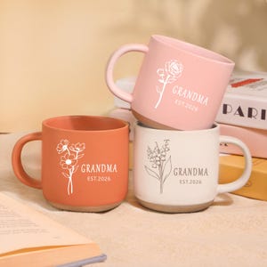 Peut inclure: Trois mugs en c&eacute;ramique corail, rose et blanc, chacun avec un motif floral et l'inscription "GRANDMA" et "EST.2026". Les mugs ont un design simple et moderne avec une forme arrondie et une anse.