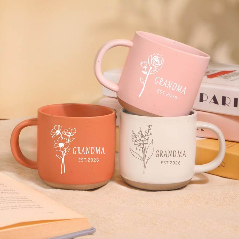 Peut inclure: Trois mugs en c&eacute;ramique corail, rose et blanc, chacun avec un motif floral et l'inscription "GRANDMA" et "EST.2026". Les mugs ont un design simple et moderne avec une forme arrondie et une anse.