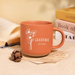 Peut inclure: Mug en c&eacute;ramique couleur terre cuite avec un motif floral blanc et le mot "GRANDMA" imprim&eacute;. Le mug a une base marron clair et une anse incurv&eacute;e. Le texte "EST.2026" est &eacute;galement imprim&eacute; sur le mug.