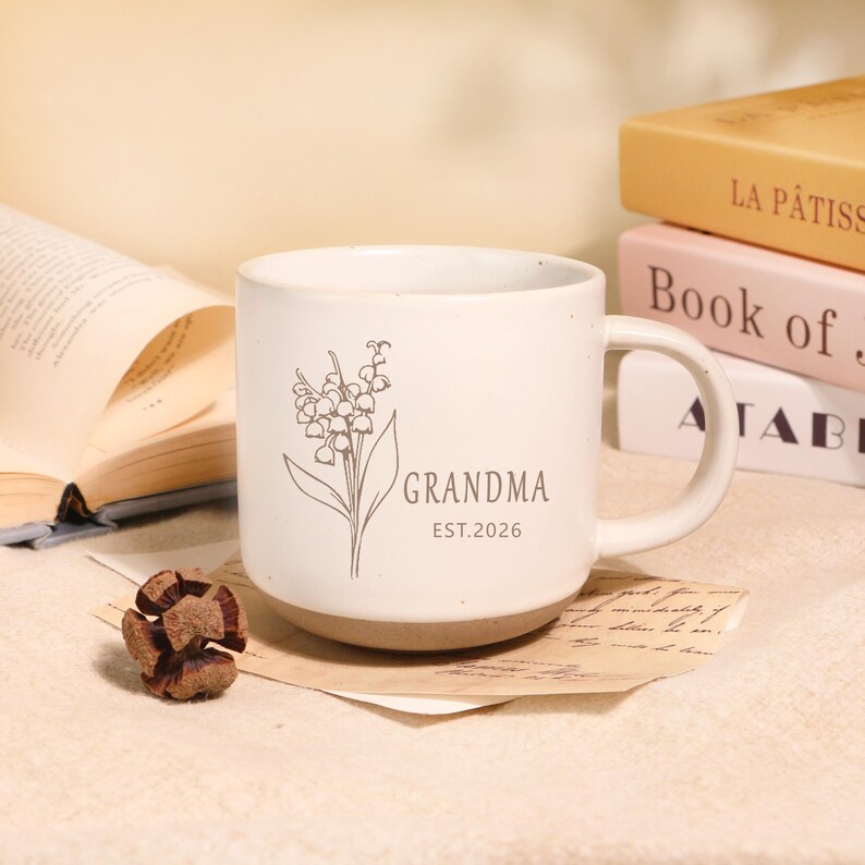 Peut inclure: Mug en c&eacute;ramique cr&egrave;me avec une base marron, orn&eacute; d'une illustration florale et du mot "GRANDMA" avec "EST.2026" en dessous. La tasse a une anse incurv&eacute;e et repose sur un morceau de papier. Des livres sont empil&eacute;s en arri&egrave;re-plan.