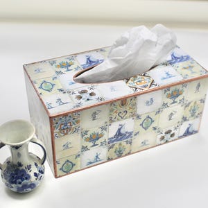 May include: A rectangular tissue box with a decorative tile pattern in blue, white, and yellow. The box has a copper-colored trim and a small, matching decorative tile near the tissue opening. A small, blue and white vase is next to the box.