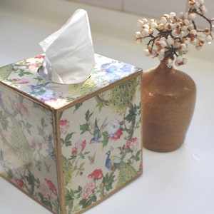 May include: A square tissue box cover with a white tissue inside. The cover is made of wood and has a floral design with peacocks and gold accents. The box is sitting on a white surface.