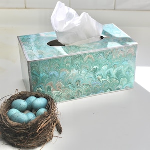 May include: A teal and white marbled tissue box with a white tissue dispenser. A nest of blue bird eggs sits in front of the tissue box.