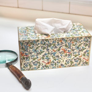 May include: A white tissue box cover with a floral pattern in shades of blue, green, and orange. The cover has a gold trim and is made of a sturdy material.