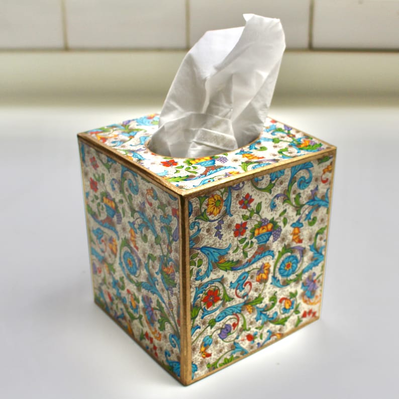 May include: A white tissue box cover with a colourful floral pattern. The pattern features blue, green, yellow, red, and white flowers and leaves on a white background. The box is made of wood and has a gold trim.