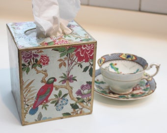 May include: A white tissue box with a floral and bird design. The box is made of wood and has a gold trim. The tissue box is sitting on a white surface.