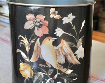 May include: A black metal waste bin with a decorative floral and bird design. The design features a bird with outstretched wings, flowers, and butterflies in peach, white, and gold. The bin has a silver rim.