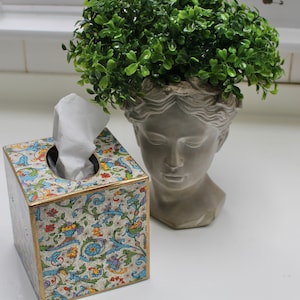 May include: A white tissue box with a colourful floral design sits on a white surface next to a grey plaster bust with a green plant growing out of the top.