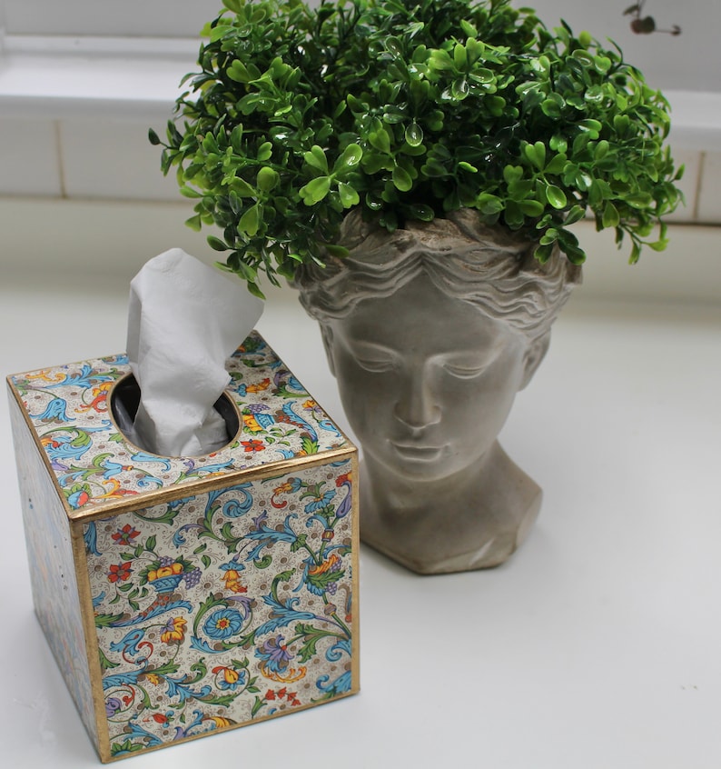 May include: A white tissue box with a colourful floral design sits on a white surface next to a grey plaster bust with a green plant growing out of the top.