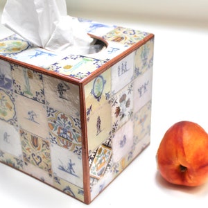 May include: A square tissue box cover with a colorful tile design. The cover is made of wood and has a clear protective coating. The tiles are in various shades of blue, yellow, and white. The box is sitting on a white surface next to a peach.