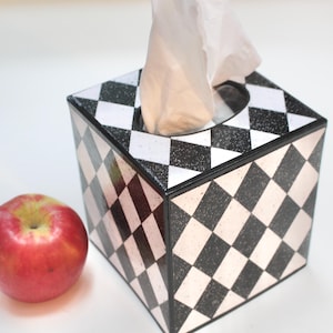 May include: A black and white checkered tissue box with a white tissue inside. The tissue box is sitting on a white surface next to a red apple.