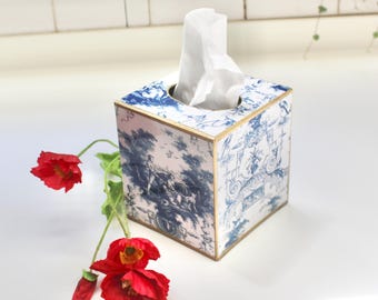 May include: A square tissue box cover featuring a blue and white toile de jouy pattern with gold-toned edges. A single white tissue protrudes from the top opening. Three bright red poppy flowers with green stems are positioned beside the box.
