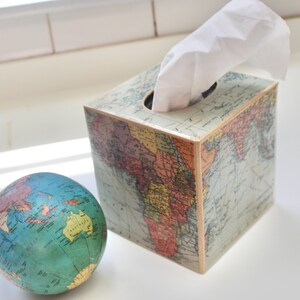 May include: A square tissue box with a world map design. The box is light-colored with a vintage map print in shades of blue, green, yellow, and red. White tissues are pulled from the top opening. A small globe is next to the box.