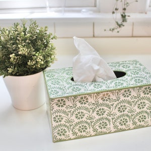 May include: A white tissue box with a green floral pattern. The box is made of wood and has a rectangular shape. The tissue box is sitting on a white surface.