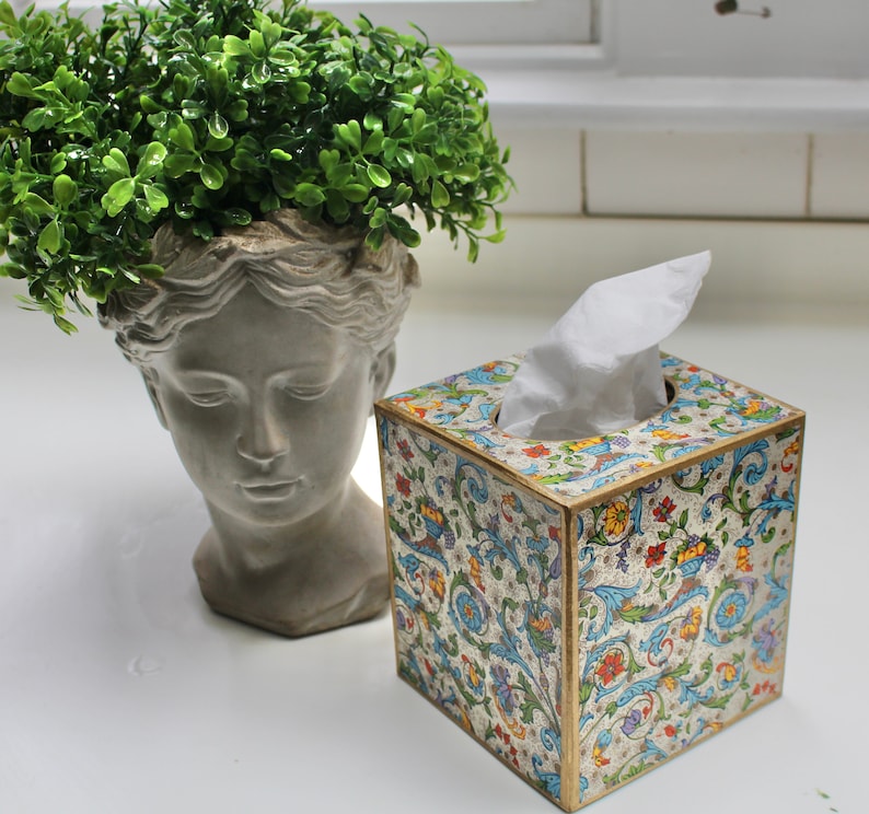 May include: A decorative tissue box cover with a floral pattern in shades of blue, green, and gold. The cover is made of wood and has a white tissue box inside.