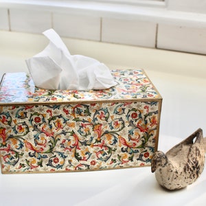 May include: A tissue box cover with a floral pattern in shades of red, green, blue, and gold. The cover is made of a hard material and has a gold trim. A white tissue is visible inside the box. A white ceramic bird figurine is in the foreground.