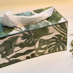 May include: A white tissue box with a green and white tropical leaf design. The box is sitting on a white surface.