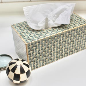 May include: A tissue box cover with a geometric pattern of green and gold leaves. The cover is made of wood and has a rectangular shape.