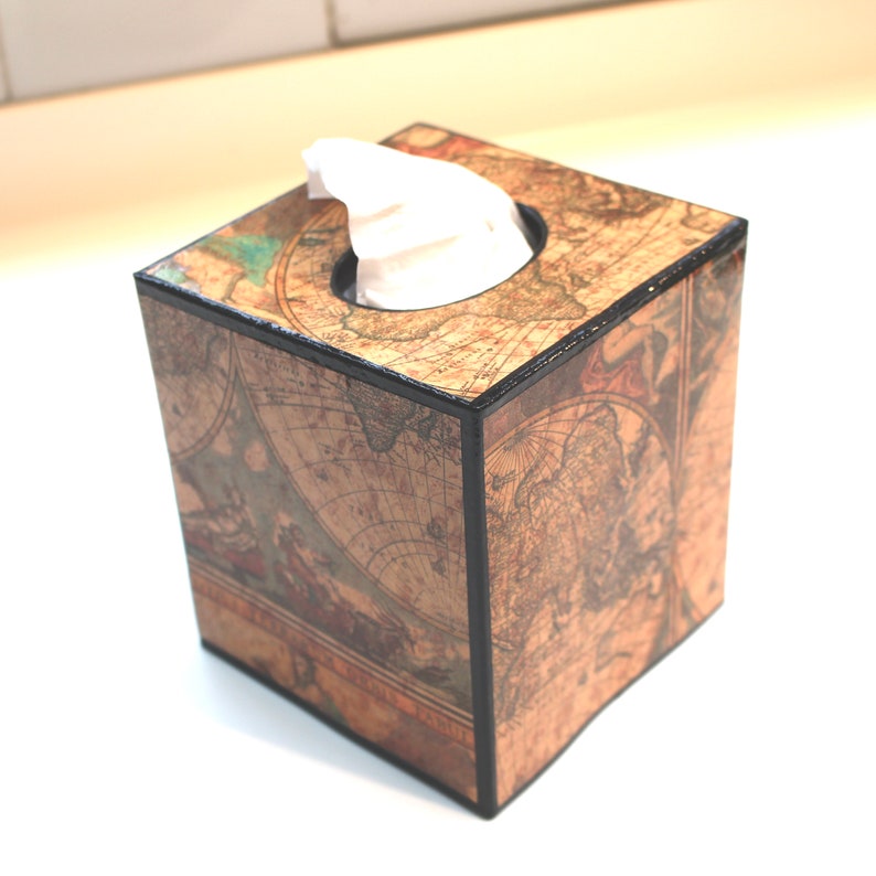 May include: A square tissue box featuring a vintage world map design in shades of brown, beige, and teal. The box has a black trim and a circular opening at the top, with white tissues visible. The box is a decorative item for home use.