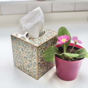 May include: A tissue box with a floral pattern and a white tissue dispenser. A pink flower pot with pink flowers is next to the tissue box.