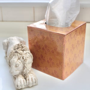 May include: A gold tissue box cover with a decorative pattern and a white tissue box inside. A white ceramic lion figurine is in the foreground.