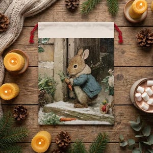 May include: A drawstring gift bag featuring a classic illustration of Peter Rabbit holding a carrot, wearing a blue jacket. The bag has a cream-colored top and red drawstrings. The scene is set in a snowy garden, evoking a cozy, winter theme.