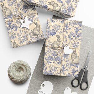 Puede incluir: Varios regalos envueltos con papel de regalo con estampado floral y conejos azules y beige. Cada regalo está atado con hilo y tiene una etiqueta blanca. También se ven una madeja de hilo, unas tijeras y etiquetas en blanco.