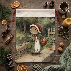 May include: A canvas drawstring bag featuring a charming illustration of a rabbit in a red hat and green coat, carrying a basket of apples and a carrot. The artwork is set in a scenic outdoor environment, evoking a storybook feel. The bag has a cream-colored top and red drawstrings.