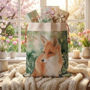 May include: A large canvas gift bag featuring a watercolor fox illustration, filled with wrapped presents. The bag has a drawstring closure with red cords. The gifts are wrapped in pastel paper and tied with ribbons. The scene is set near a window.