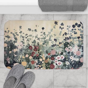 May include: A rectangular bath mat with a floral design in shades of pink, red, green, and blue. The mat has a cream-colored background and is placed on a tiled floor. A pair of gray slippers are in the foreground.
