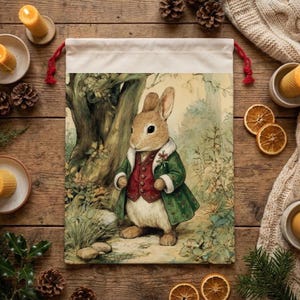 May include: A canvas gift bag featuring a watercolor illustration of a rabbit in a green coat and red vest standing in a forest. The bag has a cream-colored top with red drawstring cords. The background includes candles, pinecones, and dried orange slices.