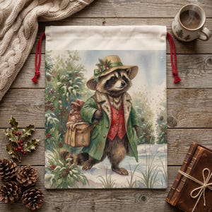 May include: A drawstring bag featuring a watercolor illustration of a raccoon in a green coat and hat, carrying a satchel. The raccoon is walking through a snowy winter scene. The bag has a cream-colored top and red drawstring.