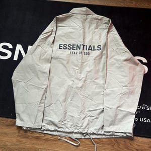 Op de afbeelding: Lichtbeige coachjack met de tekst "ESSENTIALS FEAR OF GOD" in zwart op de rug gedrukt. De jas heeft een trekkoord in de zoom en een kraag. De jas ligt plat op een donkere ondergrond.