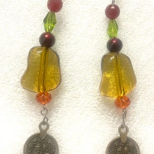 Puede incluir: Un par de pendientes colgantes con una estética vintage. Cada pendiente presenta un dije de moneda plateada, un colgante grande de vidrio color ámbar y varias cuentas en rojo, verde y naranja. Los pendientes están suspendidos de ganchos plateados.