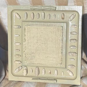 May include: A square, light green frame with a central square of beige fabric. The frame is decorated with small, oblong, beige-colored embellishments around the perimeter. The background is a striped beige and white fabric.