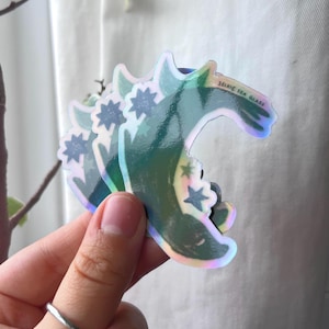 以下が含まれることがあります： 星のアクセントが付いた、ホログラフィックの三日月型ステッカー。ステッカーは、色のスペクトルを反射する虹色の仕上がりになっています。「SELKIE SEA GLASS」の文字がステッカーに印刷されています。ステッカーは手で持たれています。